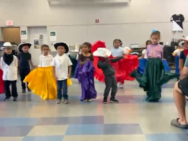 Students performing a group dance in colorful skirts and hats.